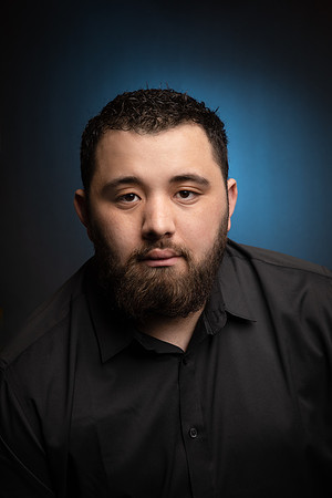 A young man poses for a headshot against a dark background with blue light inside the studio.